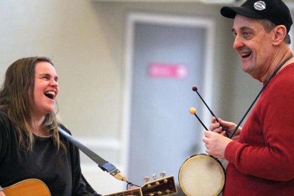 A woman playing guitar smiles at a man in a black hat playing a drum
