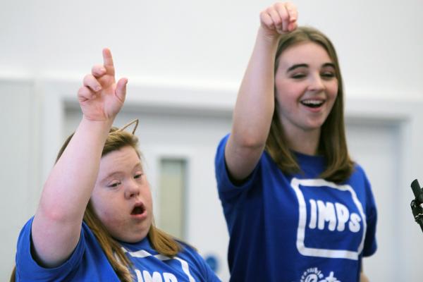Two people sing and sign on stage wearing blue IMPs t-shirts