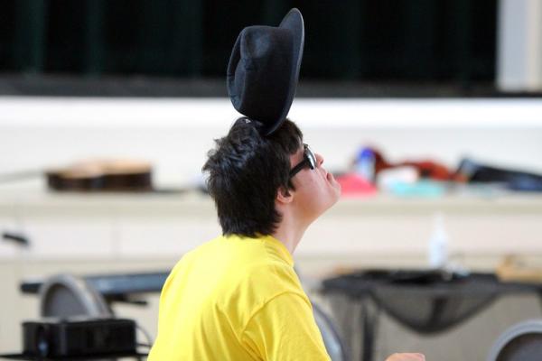 A person in a yellow top balances a black hat on his head 