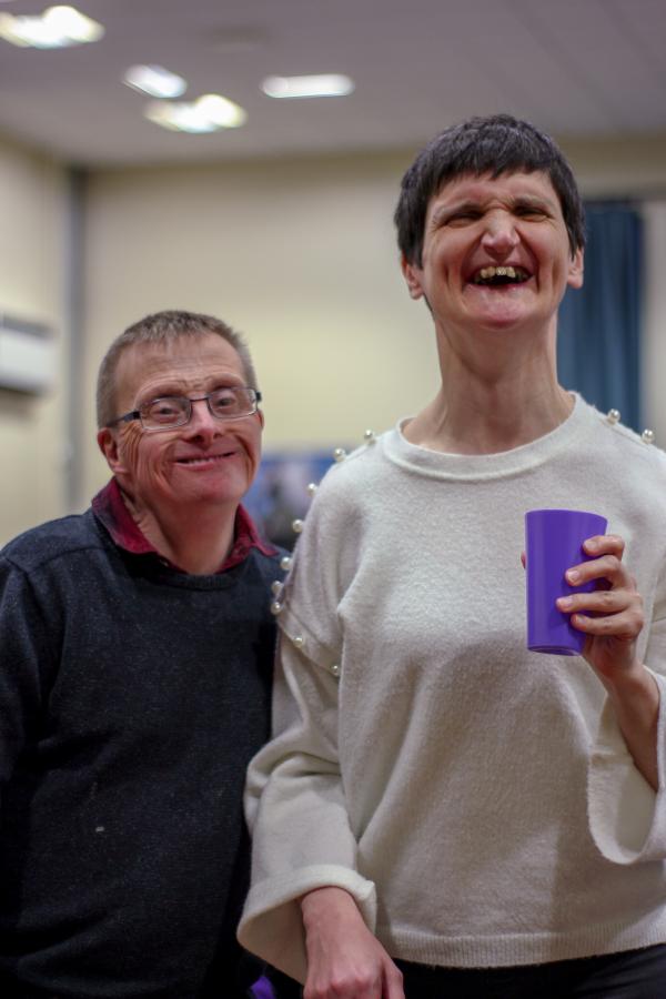Two people stand side by side smiling. One is holding a purple cup