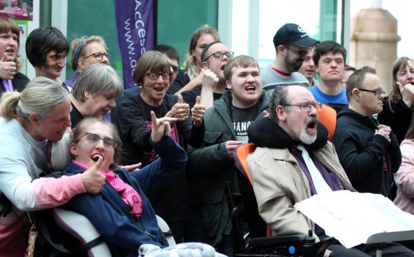 A group of people sing together. A lady in the centre stands with both thumbs up