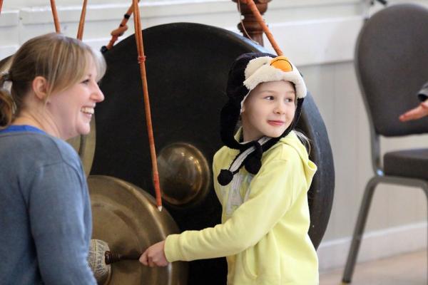 A girl wearing a penguin hat smiles and plays a large Gamelan with a mallet 