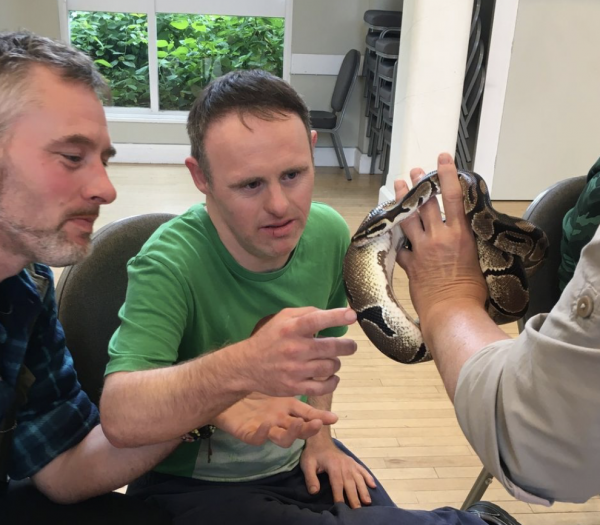 A man wearing a green t-shirt softly strokes a snake that is being held by Mags